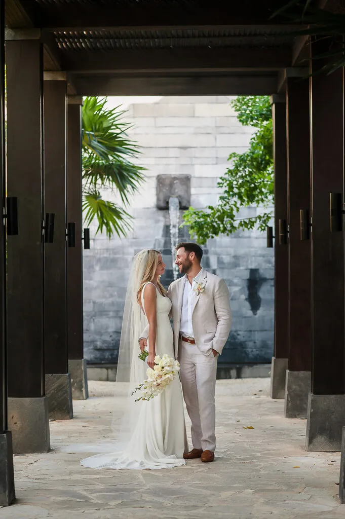 Bride and groom in elegant white attire pose at luxury Riviera Maya resort with tropical palms and modern architecture