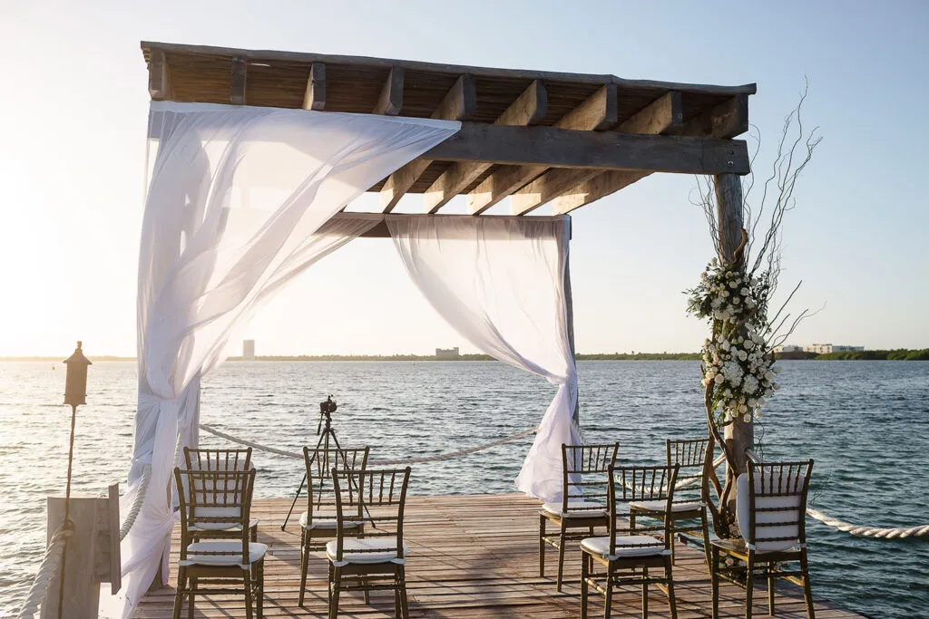 Waterfront wedding ceremony setup with wooden pergola and white draping in Cancun Riviera Maya Mexico