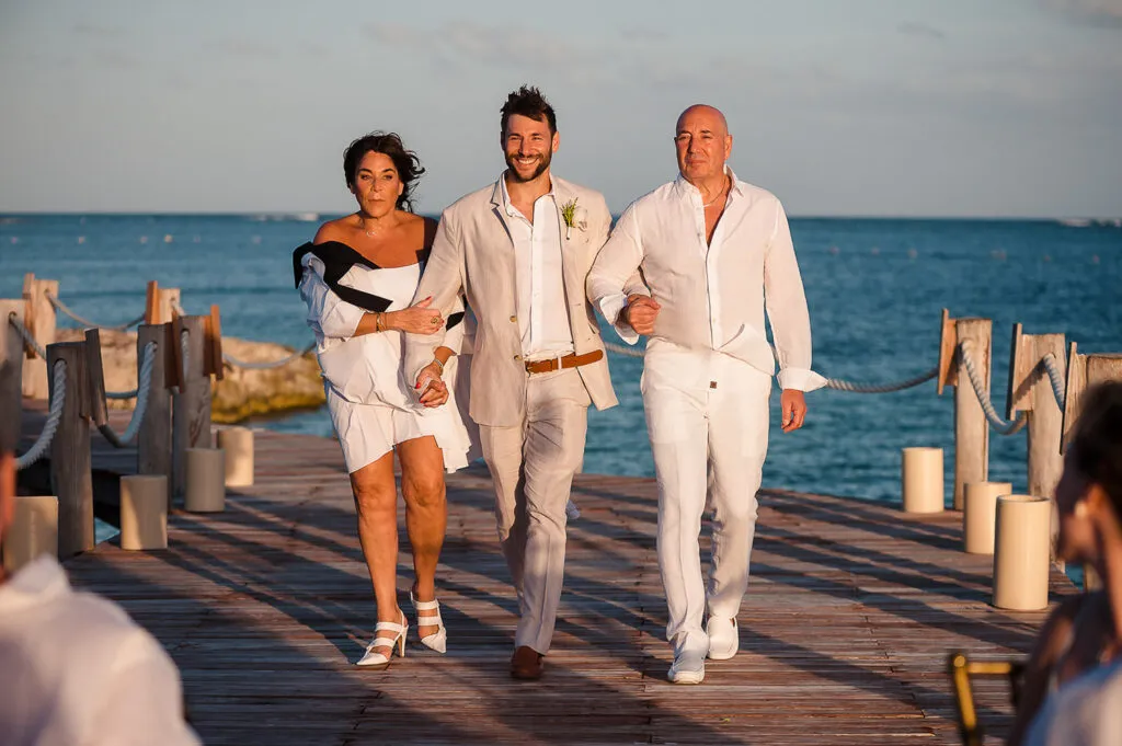 Groom walking down wooden dock with parents during beach wedding ceremony in Cancun Riviera Maya Mexico