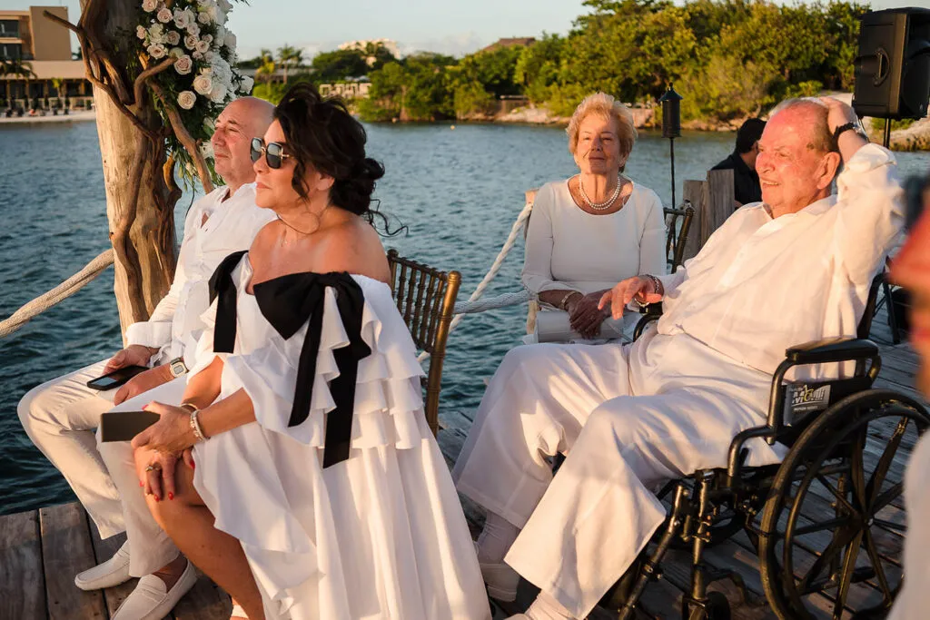 Waterfront wedding ceremony in Cancun with couple and family celebrating at sunset near lagoon with floral arch