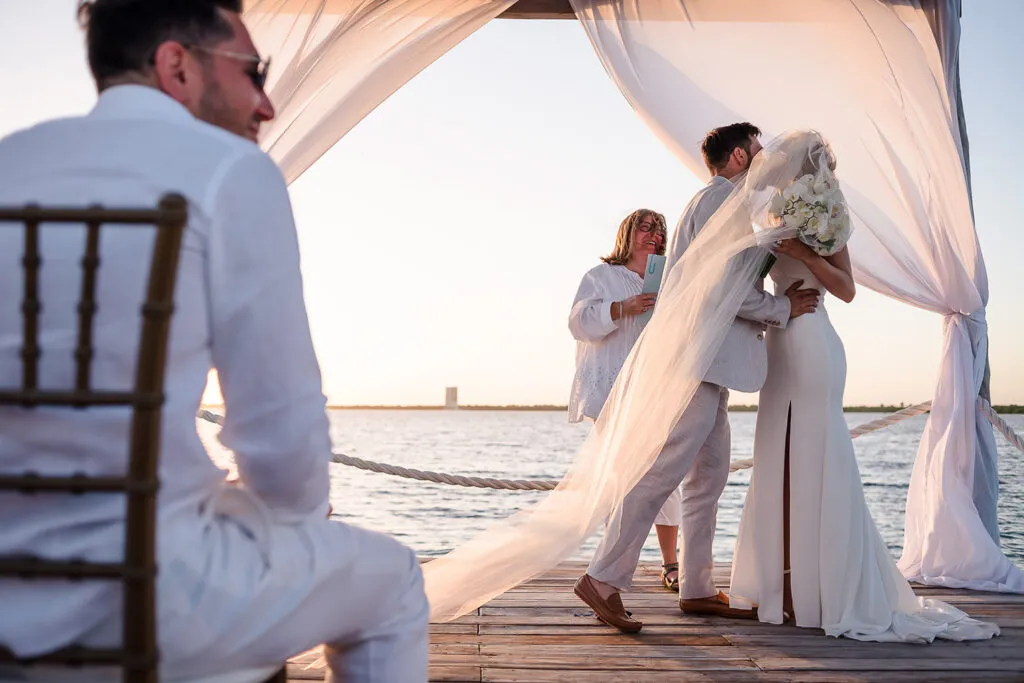 Sunset beach wedding ceremony in Cancun with couple kissing under white fabric arch on wooden dock over Caribbean waters