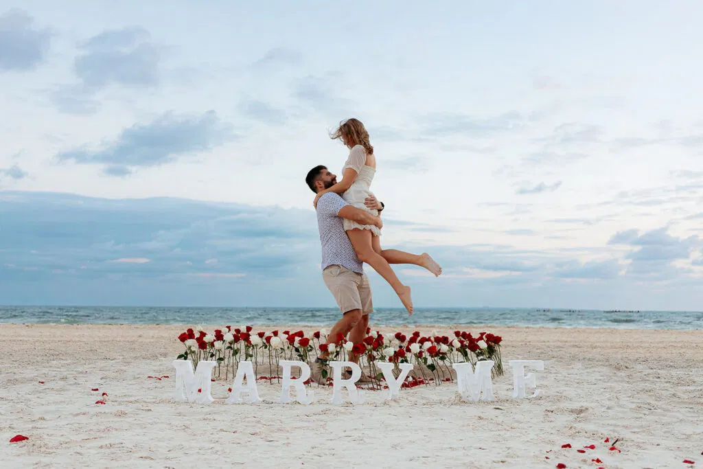 Beach proposal in Cancun with couple celebrating engagement surrounded by red roses and MARRY ME sign on white sand