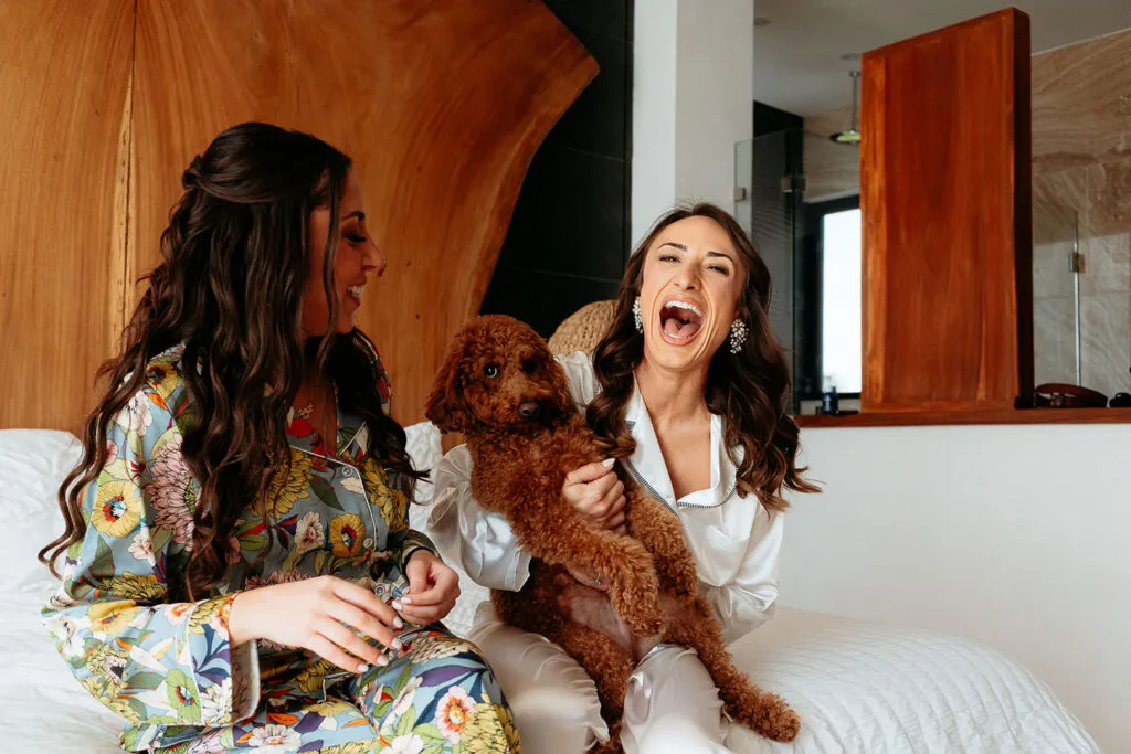 Two brides laughing with poodle dog during wedding preparation in luxury Cancun resort suite, Riviera Maya wedding photography