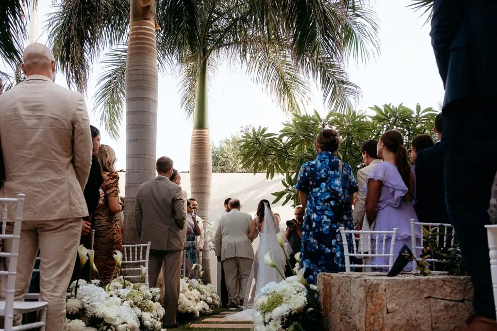 Tropical outdoor wedding ceremony in Cancun with palm trees, white flowers, and guests watching bride and groom exchange vows