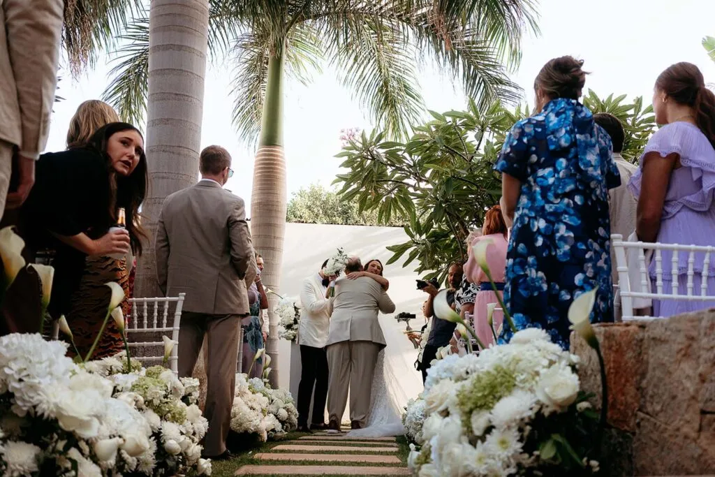 Tropical beach wedding ceremony in Cancun with bride and groom embracing surrounded by white flowers and palm trees