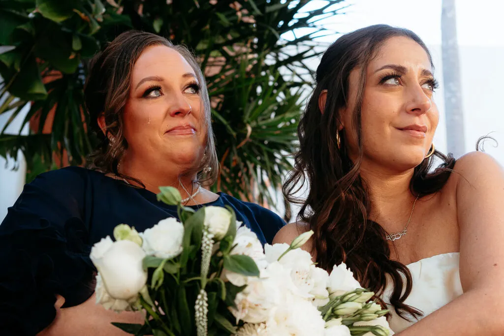 Two brides embrace during emotional wedding ceremony in Cancun with tropical palms and white bouquets