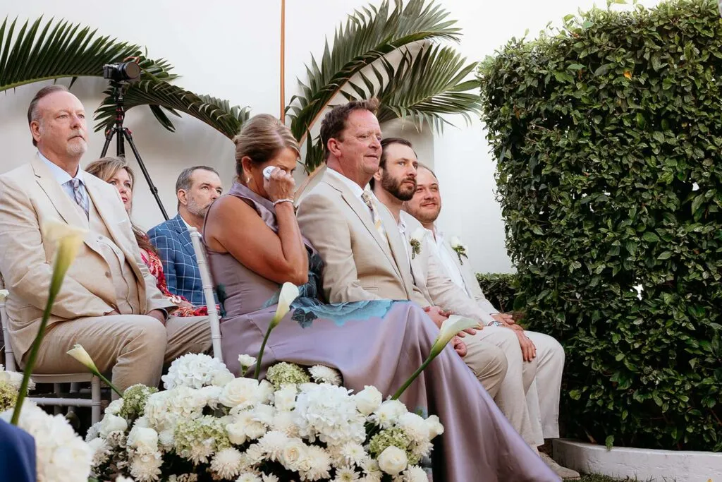 Wedding guests in light suits watching outdoor ceremony in tropical Cancun setting with palm trees and white flowers