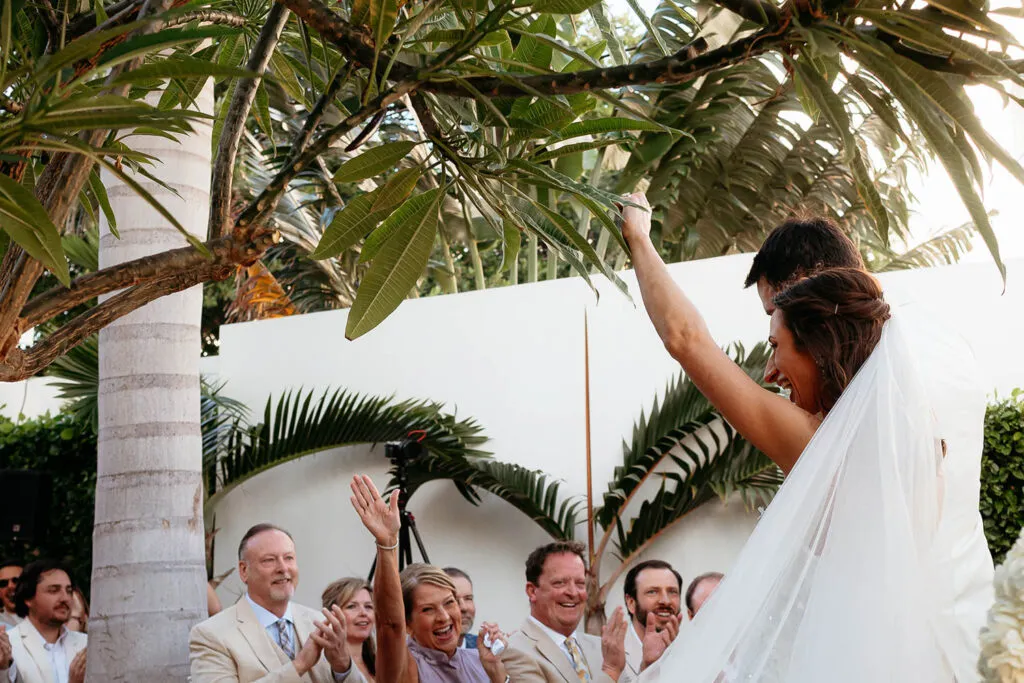 Bride celebrating at tropical Cancun wedding ceremony with palm trees and guests cheering in Riviera Maya Mexico