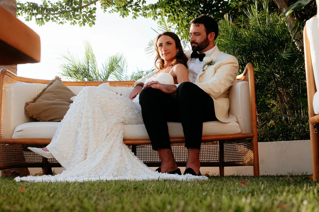Newlywed couple in elegant wedding attire relaxing on outdoor furniture at luxury Cancun resort with tropical greenery