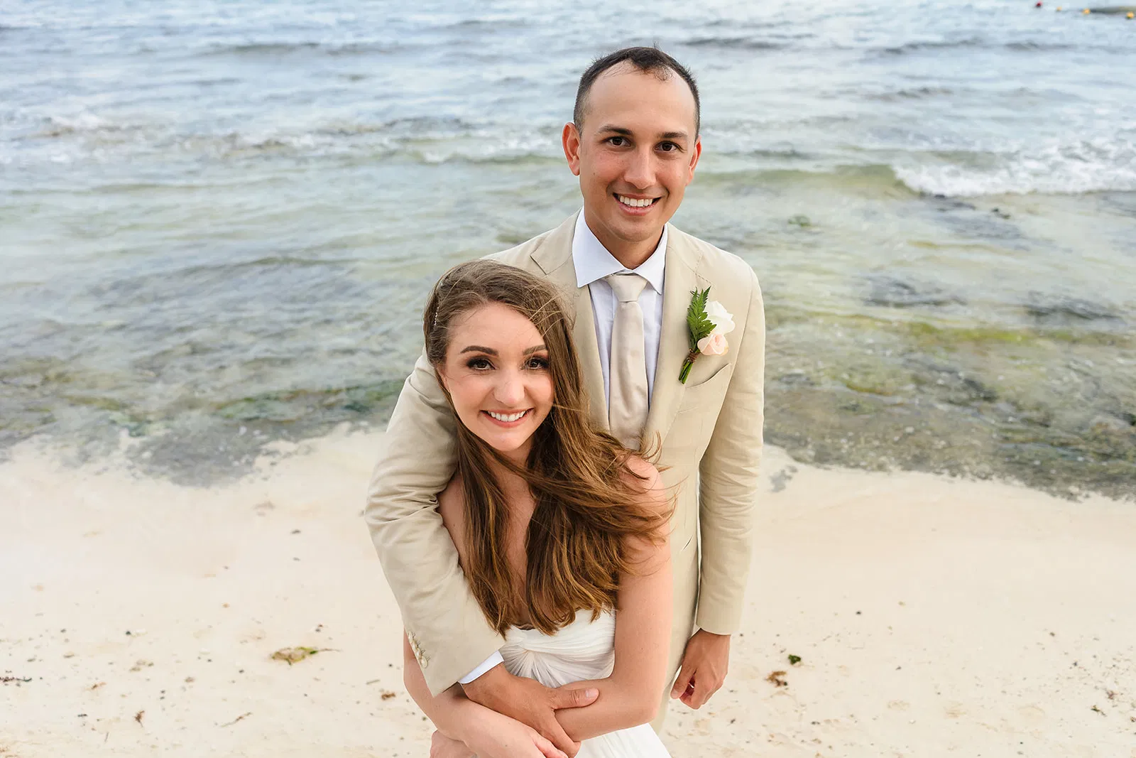 Beach wedding couple embracing on Tulum shores in elegant attire with boutonniere and flowing hair in ocean breeze