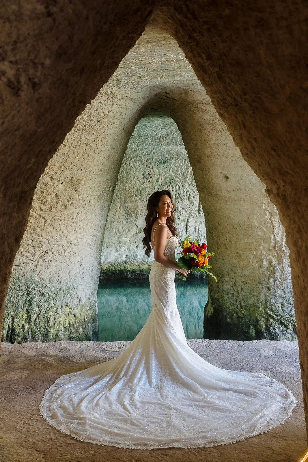 Bride holding colorful bouquet framed by stone archway at Hotel Xcaret destination wedding in Riviera Maya