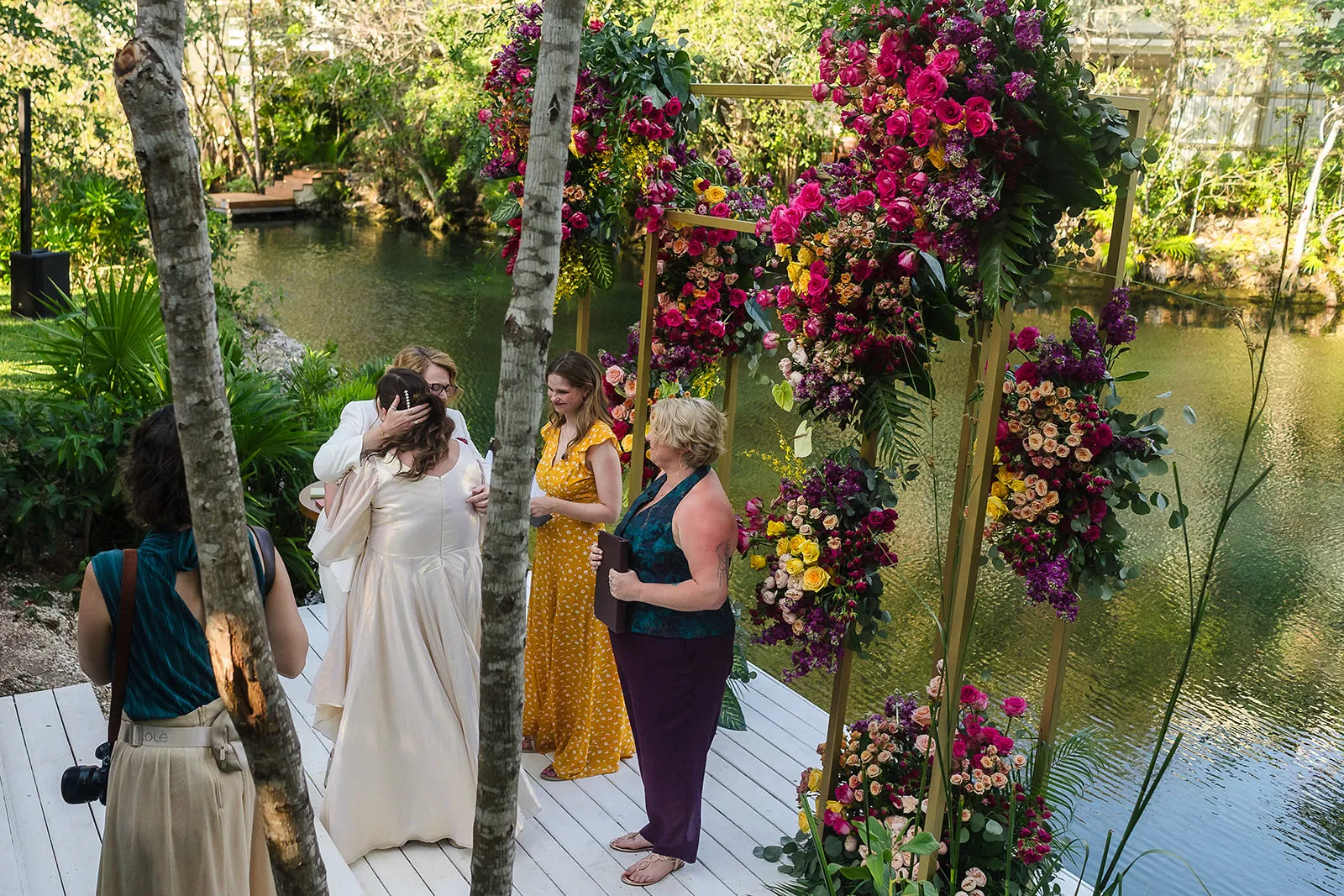 LGBTQ wedding ceremony at Rosewood Mayakoba with same-sex couple exchanging vows under vibrant floral arch by cenote waters