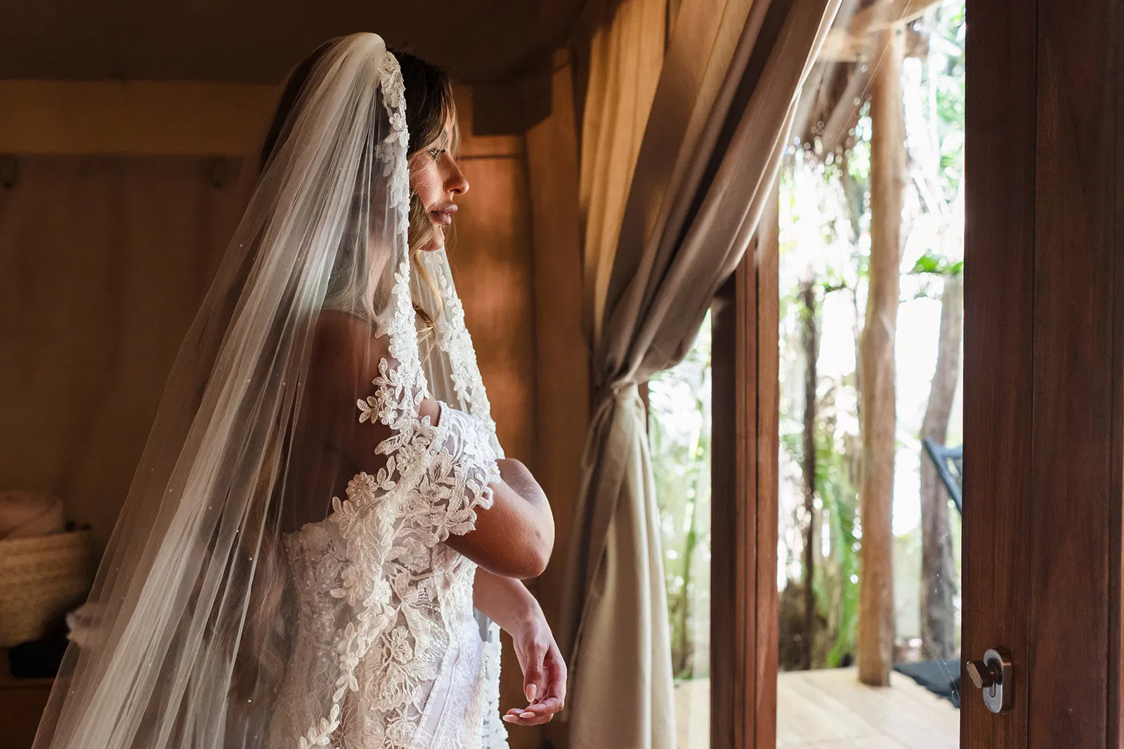 Bride in lace wedding dress and veil looking out window at tropical Tulum wedding venue in Mexico