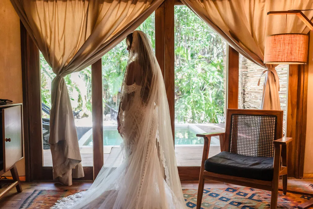 Bride in lace wedding dress and veil looking out window at tropical Tulum resort before destination wedding ceremony