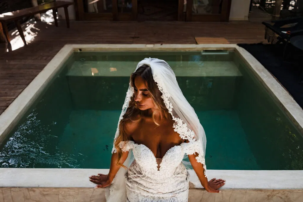 Bride in white lace wedding dress and veil sitting by turquoise pool in Tulum Mexico destination wedding photography