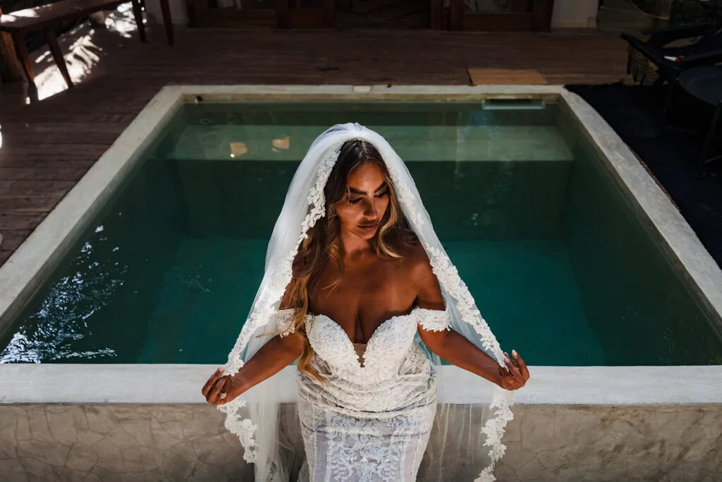 Bride in white lace wedding dress and veil posing by turquoise pool in Tulum Mexico destination wedding photography