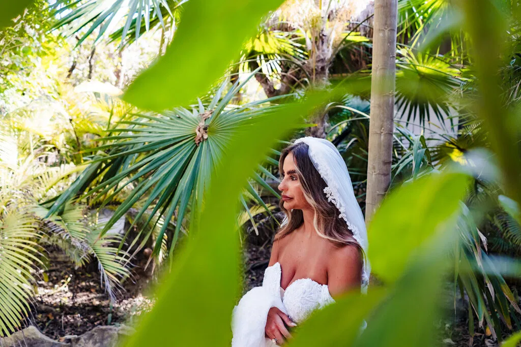Bride in white wedding dress and veil posing among tropical palm trees in Tulum Mexico for destination wedding photography