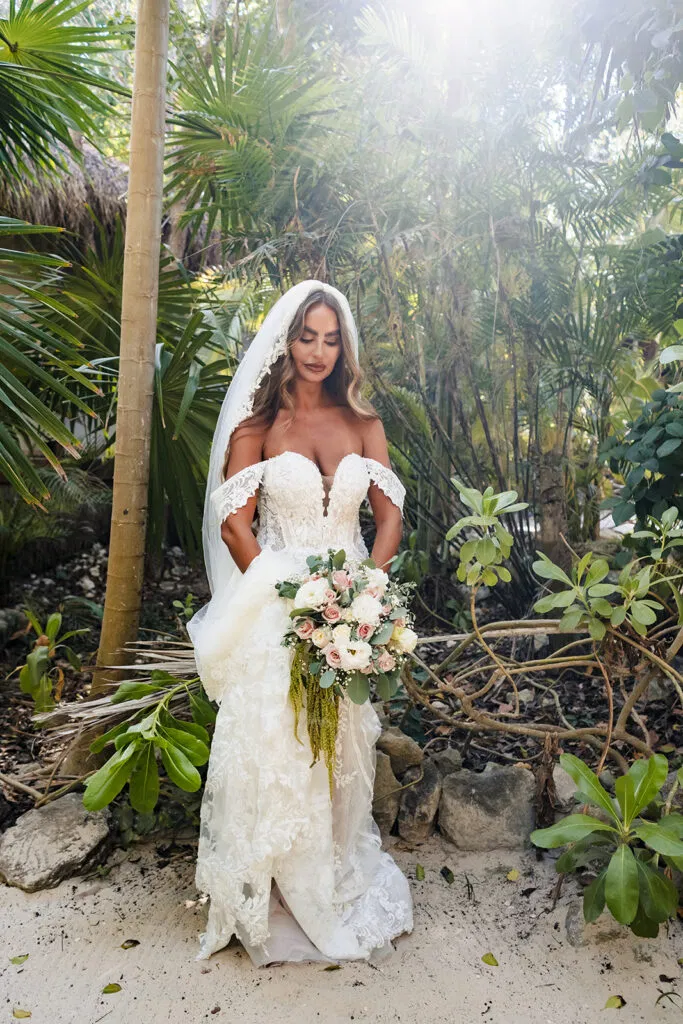 Bride in lace wedding dress with bouquet posing in tropical jungle setting for destination wedding photography in Tulum Mexico