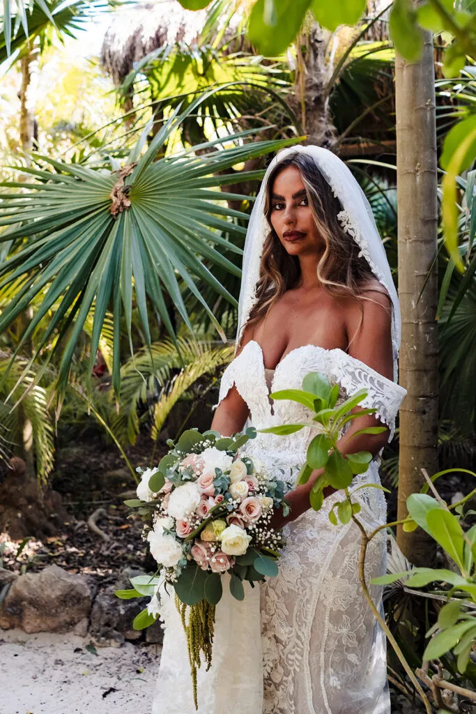 Bride in white lace dress holding bouquet among tropical palms for destination wedding photography in Tulum Mexico