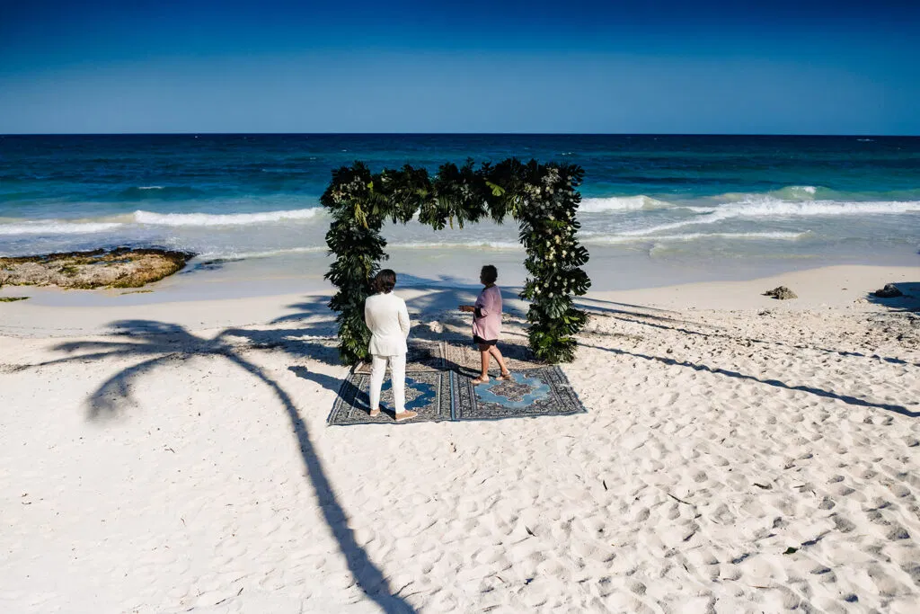 Beach wedding ceremony in Tulum Mexico with couple under tropical greenery arch on white sand near turquoise ocean waves
