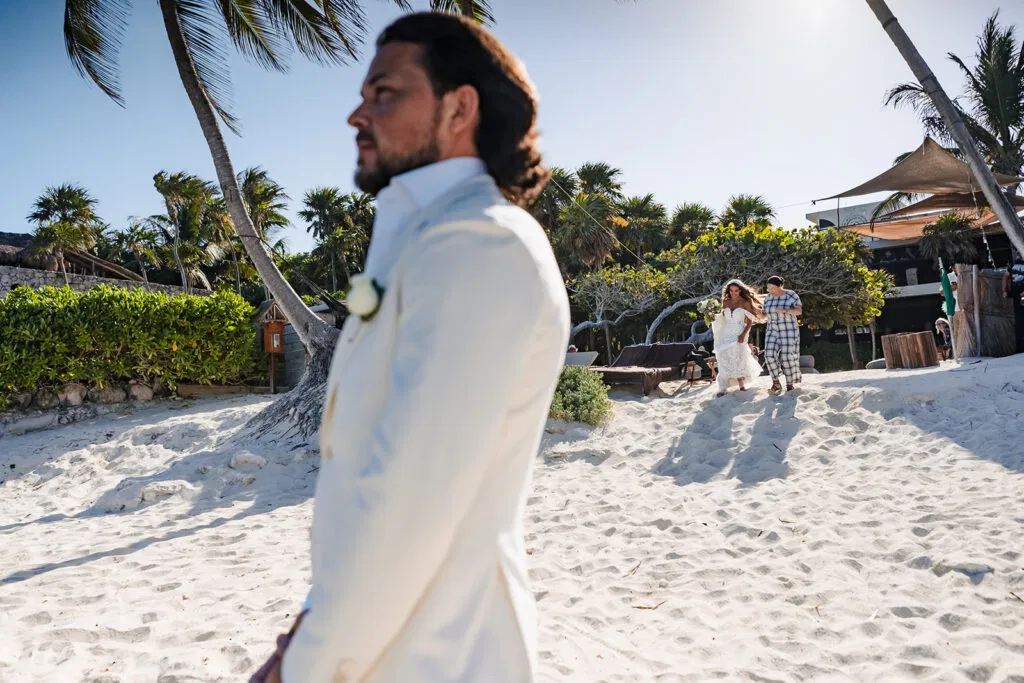 Groom in white shirt awaits bride's first look on pristine Tulum beach with palm trees for destination wedding photography