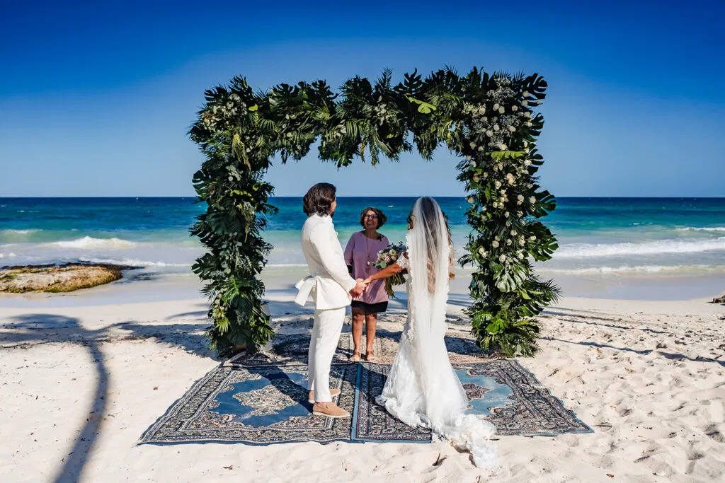 Beach wedding ceremony in Tulum Mexico with tropical floral arch, bride and groom exchanging vows on white sand beach
