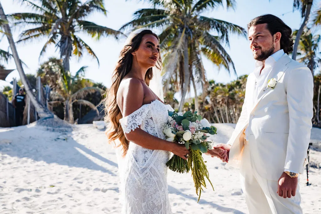 Beach wedding ceremony in Tulum Mexico with bride and groom holding hands on white sand with palm trees in background