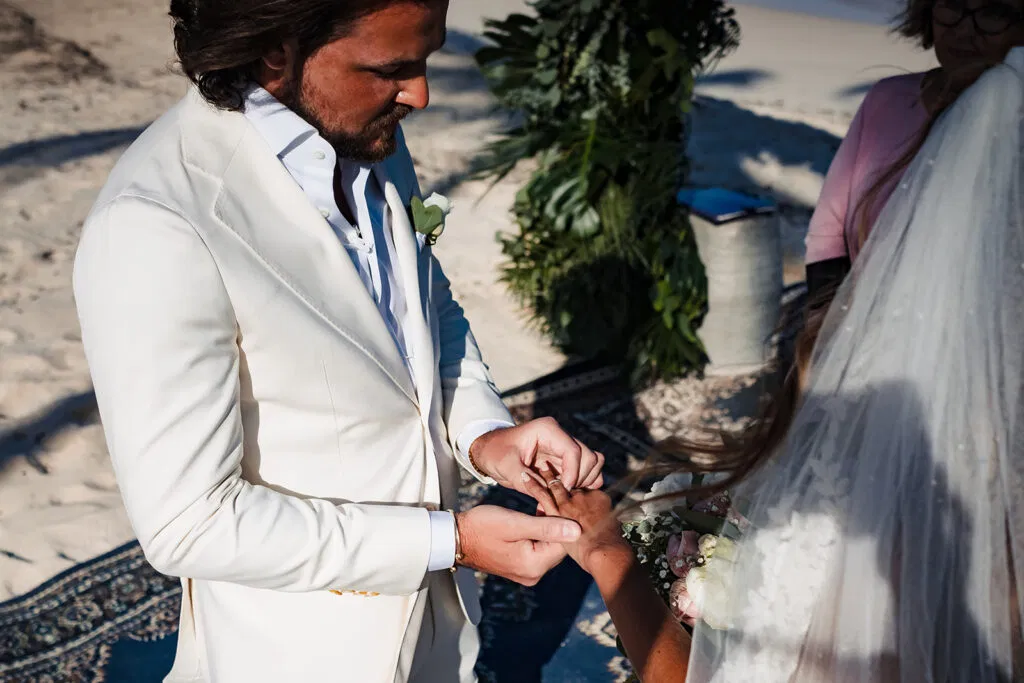 Groom in white suit placing wedding ring on bride's finger during beach wedding ceremony in Tulum Mexico