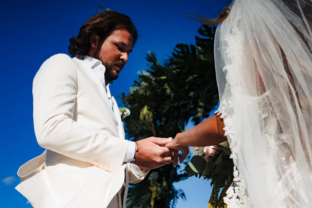 Groom placing wedding ring on bride's finger during outdoor Tulum Mexico beach wedding ceremony with palm trees and blue sky