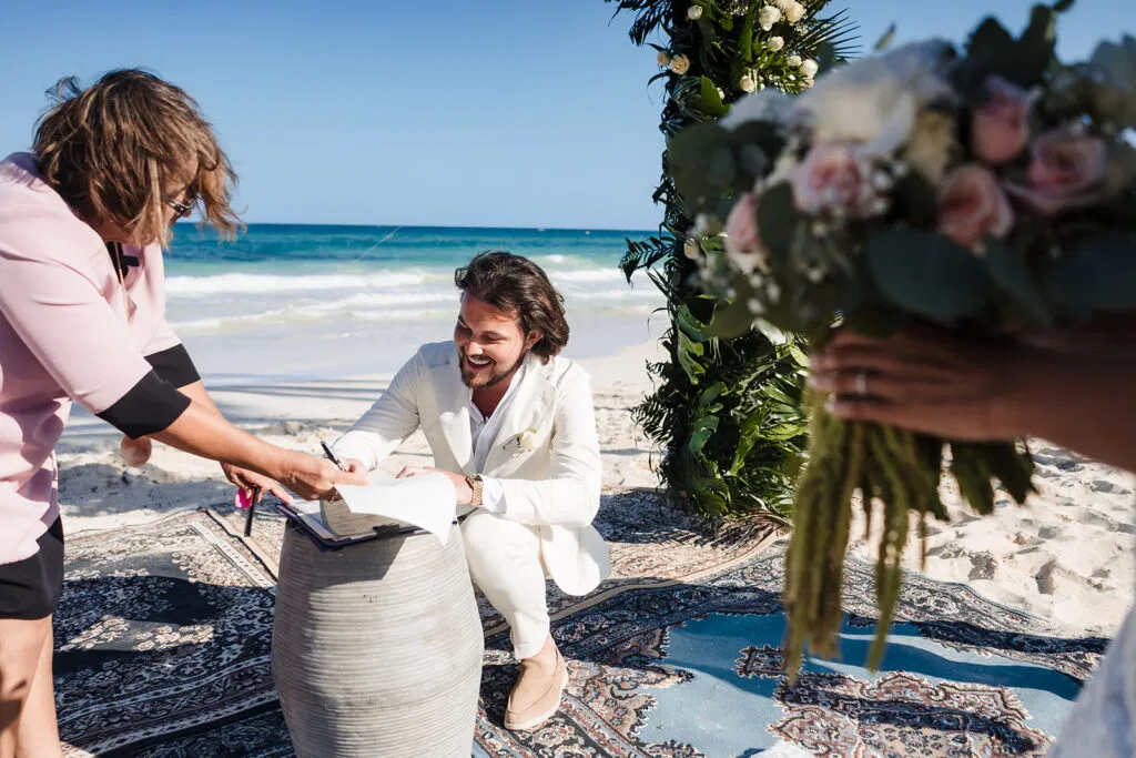 Groom signing marriage certificate at intimate beach wedding ceremony in Tulum Mexico with ocean backdrop and tropical flowers