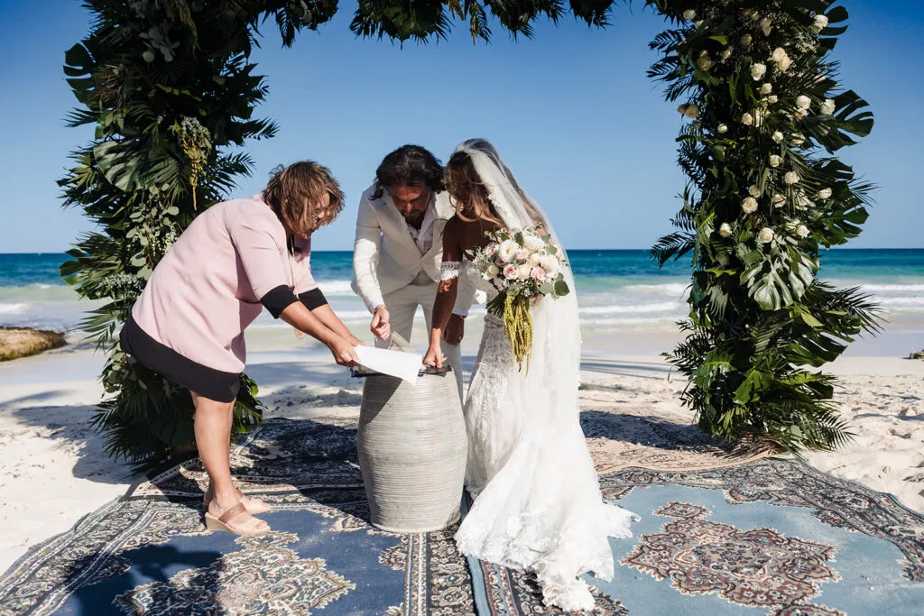 Beach wedding ceremony in Tulum with couple and officiant performing sand unity ritual under tropical floral arch