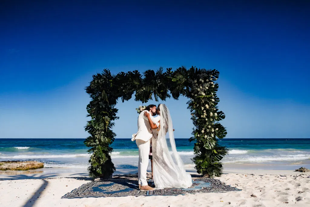 Beach wedding ceremony in Tulum Mexico with couple kissing under tropical greenery arch on white sand with blue ocean