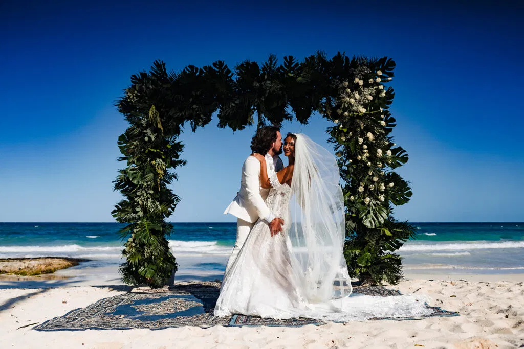 Bride and groom embrace under tropical floral arch on Tulum beach - destination wedding photography Mexico