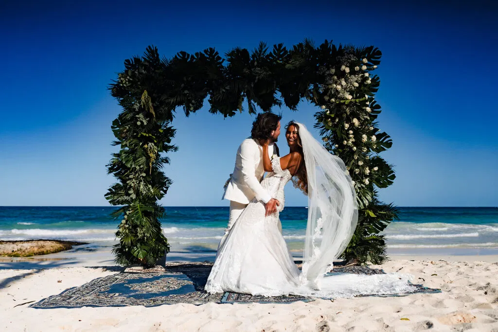 Beach wedding ceremony in Tulum Mexico with bride and groom under tropical floral arch overlooking turquoise Caribbean Sea