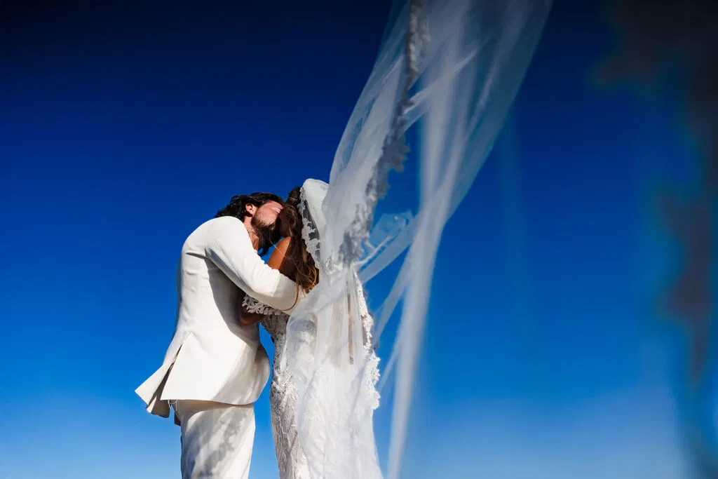 Romantic bride and groom kissing under flowing veil against clear blue Tulum sky - destination wedding photography Mexico