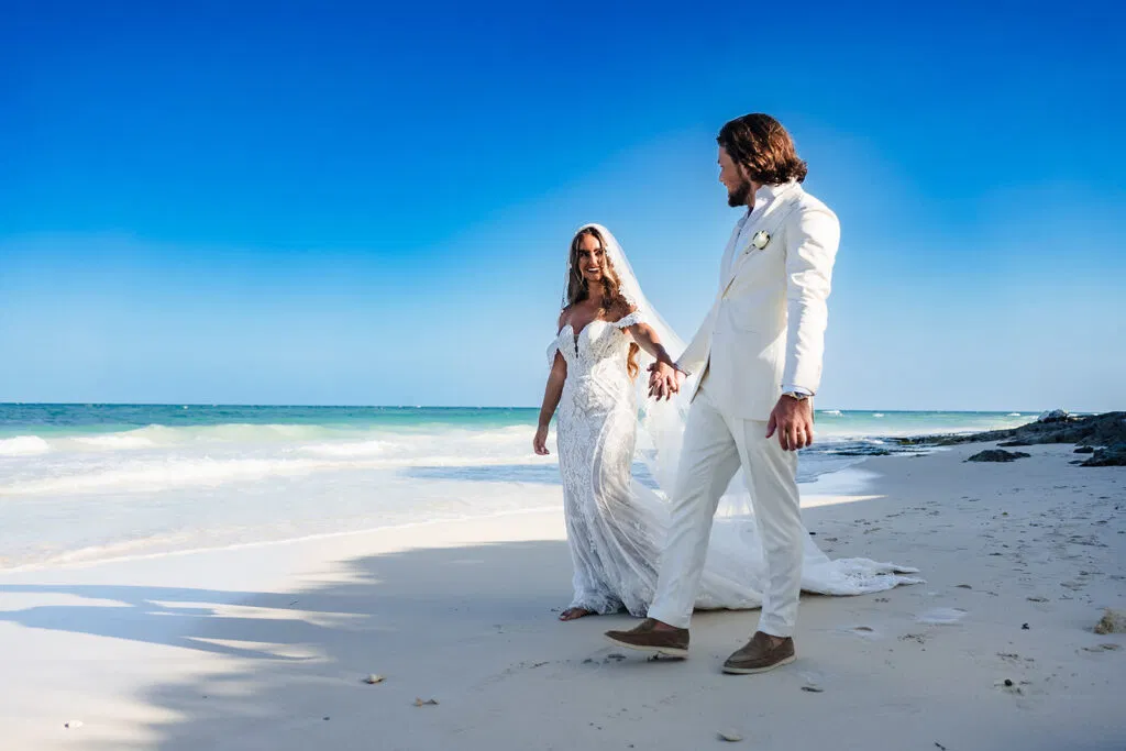 Beach wedding couple walking hand in hand on white sand in Tulum Mexico with turquoise ocean and blue sky backdrop