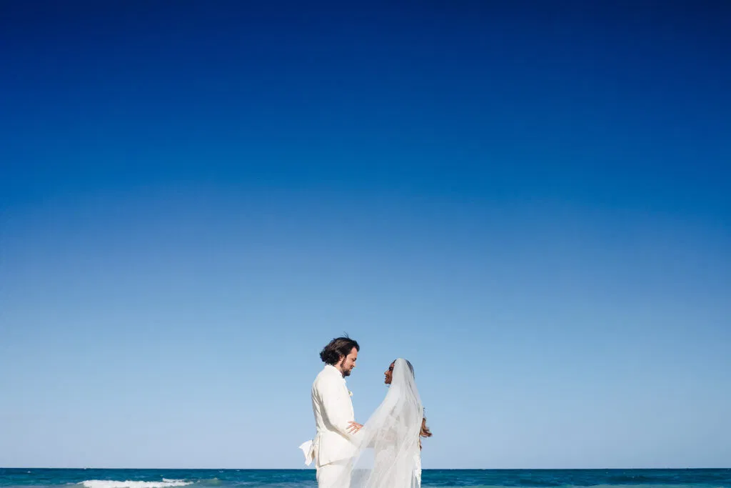 Romantic bride and groom in white flowing attire embracing on Tulum beach with crystal blue Caribbean sky backdrop