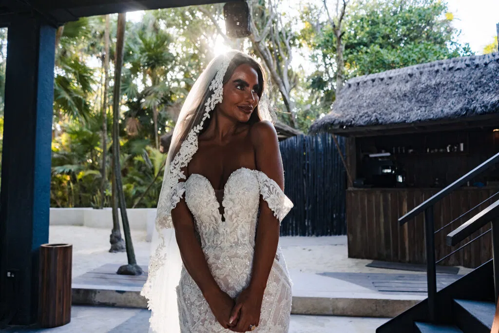 Bride in white lace wedding dress and veil at tropical Tulum resort with palm trees and thatched roof architecture