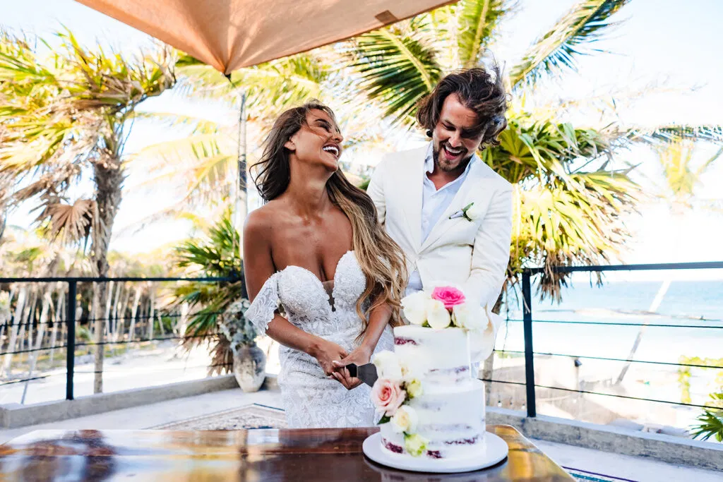 Bride and groom cutting wedding cake at tropical Tulum resort with palm trees and ocean view in Mexico