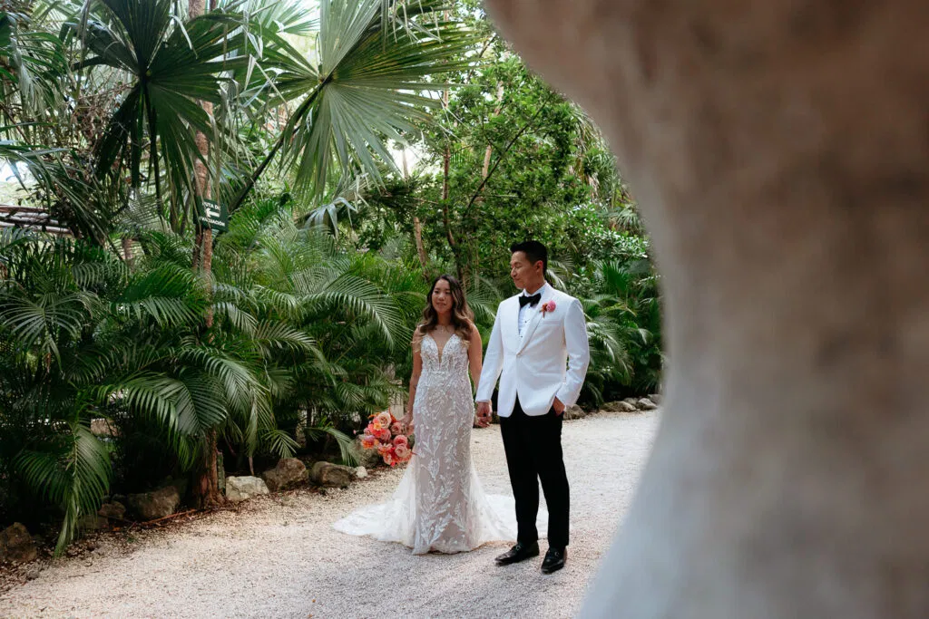 Bride and groom walking hand in hand through tropical palms at luxury Tulum Mexico wedding venue with coral bouquet