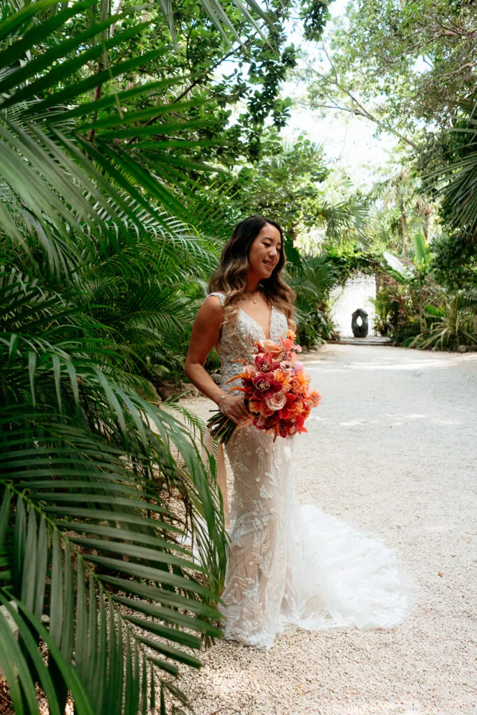 Bride in lace wedding dress holding vibrant bouquet among tropical palms in Tulum Mexico destination wedding photography