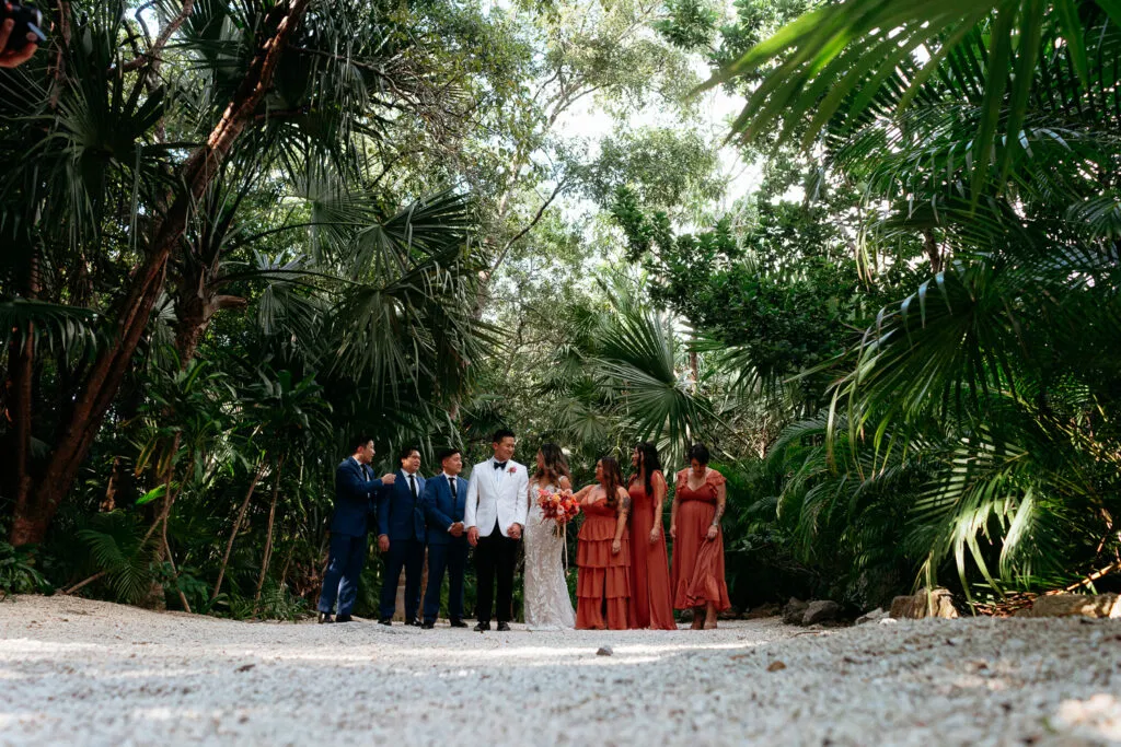 Tropical wedding party poses among lush palm trees on beach in Tulum Mexico with bride groom and coral bridesmaid dresses