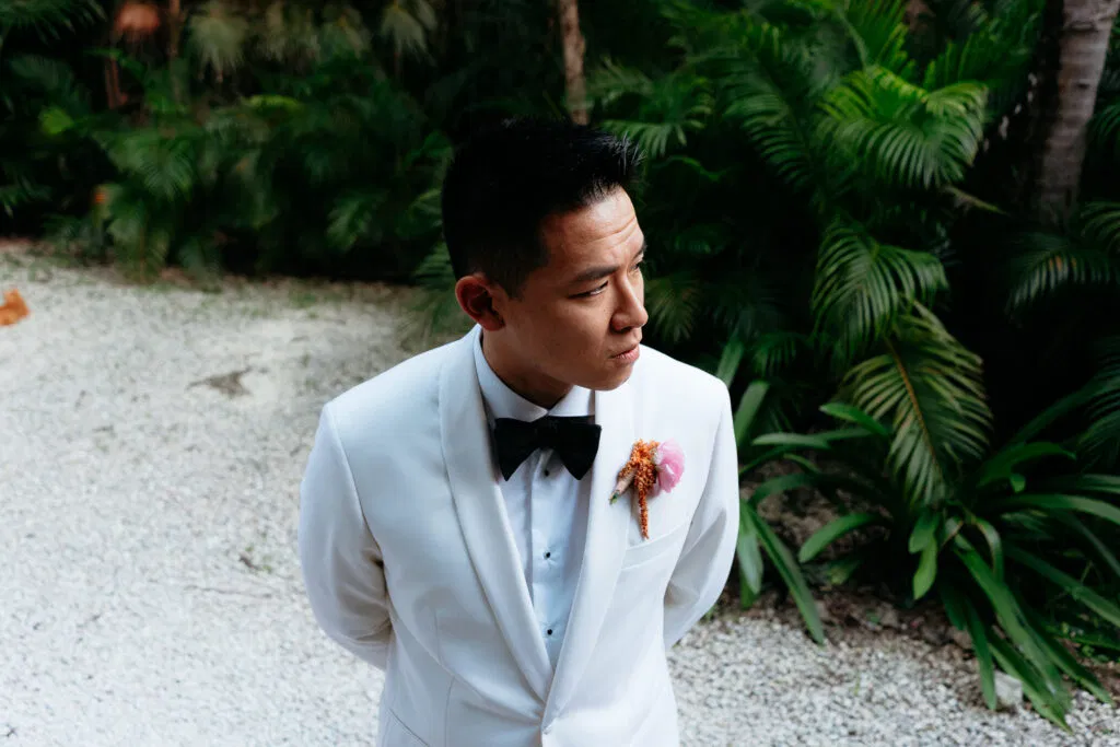 Groom in white tuxedo with floral boutonniere poses among tropical palms at luxury Tulum Mexico destination wedding