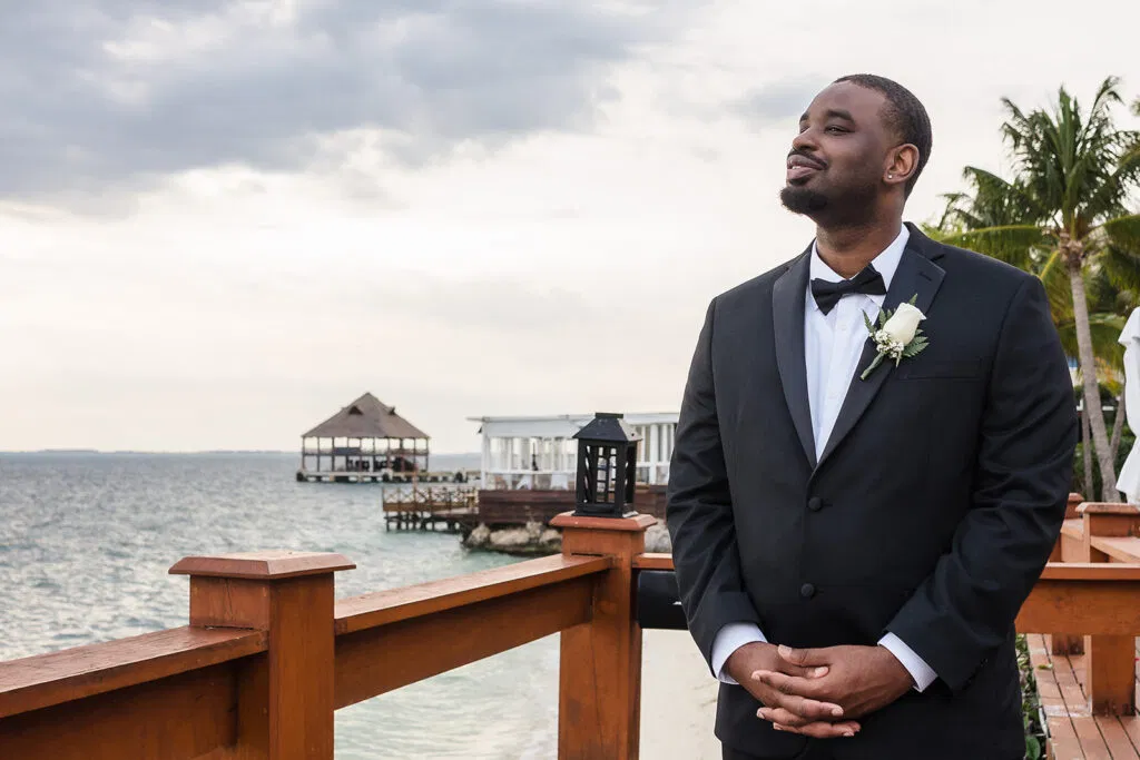 Groom in black tuxedo with white boutonniere at oceanfront Tulum wedding ceremony with tropical backdrop and pier